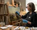 A professional art conservator wearing blue gloves and an apron meticulously cleaning an aged landscape oil painting with a fine brush in a sunlit, organized restoration studio.